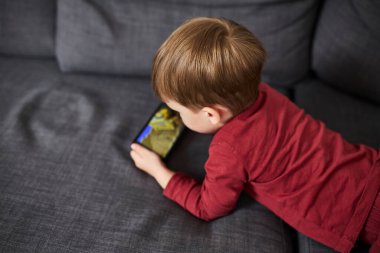 Üç yaşında bir çocuk, evinde bir kanepede oturup tablet ve akıllı telefondan çizgi film izliyor. Çocukluk kavramı, ekran zamanı, modern teknoloji ve boş zaman..