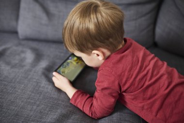 Üç yaşında bir çocuk, evinde bir kanepede oturup tablet ve akıllı telefondan çizgi film izliyor. Çocukluk kavramı, ekran zamanı, modern teknoloji ve boş zaman..