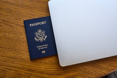 Tahta bir masada, açık bir dizüstü bilgisayar ve deri bir cüzdanın yanında yatan bir Amerikan pasaportu. Seyahat belgeleri ve kişisel aksesuarlar düz konumda dizilmiş. İş seyahati, uluslararası seyahat hazırlığı, kimlik ve güvenlik kavramı.