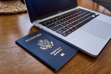 Tahta bir masada, açık bir dizüstü bilgisayar ve deri bir cüzdanın yanında yatan bir Amerikan pasaportu. Seyahat belgeleri ve kişisel aksesuarlar düz konumda dizilmiş. İş seyahati, uluslararası seyahat hazırlığı, kimlik ve güvenlik kavramı.