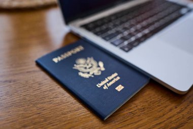 Tahta bir masada, açık bir dizüstü bilgisayar ve deri bir cüzdanın yanında yatan bir Amerikan pasaportu. Seyahat belgeleri ve kişisel aksesuarlar düz konumda dizilmiş. İş seyahati, uluslararası seyahat hazırlığı, kimlik ve güvenlik kavramı.