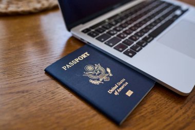 Tahta bir masada, açık bir dizüstü bilgisayar ve deri bir cüzdanın yanında yatan bir Amerikan pasaportu. Seyahat belgeleri ve kişisel aksesuarlar düz konumda dizilmiş. İş seyahati, uluslararası seyahat hazırlığı, kimlik ve güvenlik kavramı.