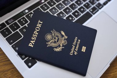 Tahta bir masada, açık bir dizüstü bilgisayar ve deri bir cüzdanın yanında yatan bir Amerikan pasaportu. Seyahat belgeleri ve kişisel aksesuarlar düz konumda dizilmiş. İş seyahati, uluslararası seyahat hazırlığı, kimlik ve güvenlik kavramı.