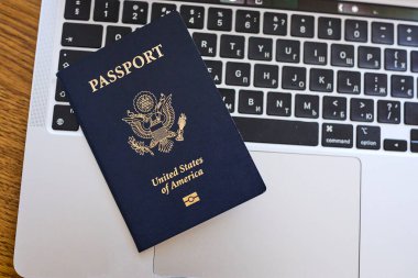 Tahta bir masada, açık bir dizüstü bilgisayar ve deri bir cüzdanın yanında yatan bir Amerikan pasaportu. Seyahat belgeleri ve kişisel aksesuarlar düz konumda dizilmiş. İş seyahati, uluslararası seyahat hazırlığı, kimlik ve güvenlik kavramı.