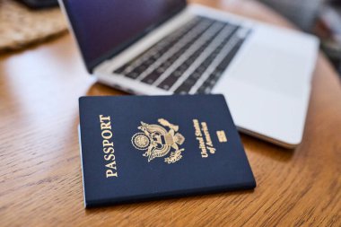 Tahta bir masada, açık bir dizüstü bilgisayar ve deri bir cüzdanın yanında yatan bir Amerikan pasaportu. Seyahat belgeleri ve kişisel aksesuarlar düz konumda dizilmiş. İş seyahati, uluslararası seyahat hazırlığı, kimlik ve güvenlik kavramı.