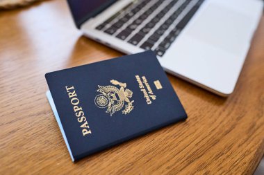 Tahta bir masada, açık bir dizüstü bilgisayar ve deri bir cüzdanın yanında yatan bir Amerikan pasaportu. Seyahat belgeleri ve kişisel aksesuarlar düz konumda dizilmiş. İş seyahati, uluslararası seyahat hazırlığı, kimlik ve güvenlik kavramı.