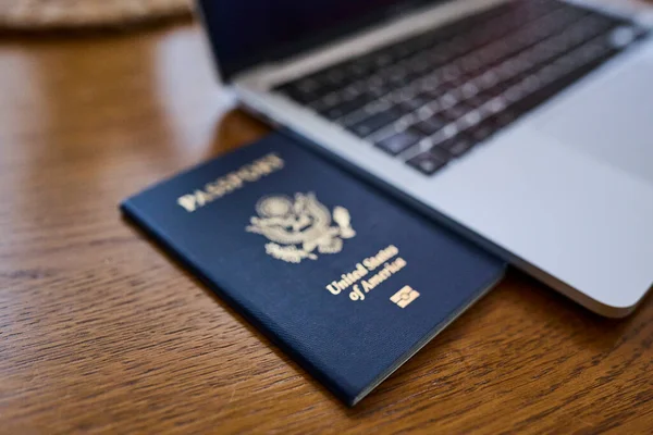 Tahta bir masada, açık bir dizüstü bilgisayar ve deri bir cüzdanın yanında yatan bir Amerikan pasaportu. Seyahat belgeleri ve kişisel aksesuarlar düz konumda dizilmiş. İş seyahati, uluslararası seyahat hazırlığı, kimlik ve güvenlik kavramı.
