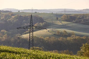 Yüksek voltajlı elektrik hatları sabah güneşinin altında Alman tepeleri boyunca uzanıyor. Enerji altyapısı, kırsal alan ve sürdürülebilir güç kavramı.