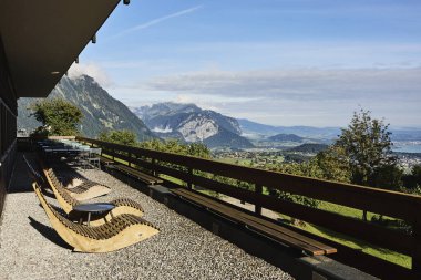Yazın İsviçre 'de Alp Dağları' na bakan bir otelin yakınındaki açık masa ve sandalyeler. Görüntü seyahat, konukseverlik, rahatlama ve açık hava yemeklerini temsil ediyor. Yaz tatili kavramı, turizm, boş zaman ve...