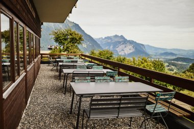 Yazın İsviçre 'de Alp Dağları' na bakan bir otelin yakınındaki açık masa ve sandalyeler. Görüntü seyahat, konukseverlik, rahatlama ve açık hava yemeklerini temsil ediyor. Yaz tatili kavramı, turizm, boş zaman ve...