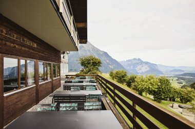 Yazın İsviçre 'de Alp Dağları' na bakan bir otelin yakınındaki açık masa ve sandalyeler. Görüntü seyahat, konukseverlik, rahatlama ve açık hava yemeklerini temsil ediyor. Yaz tatili kavramı, turizm, boş zaman ve...