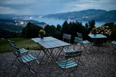 İsviçre Alpleri 'nde bir otelin yanındaki açık hava atıştırma masaları, dağlara bakan bir yer ve sıcak akşam ışığında bir göl. Görüntü rahatlamayı, seyahati, dağlık manzarayı ve akşam yemeğini temsil ediyor. Turizm kavramı, yaz tatili, konukseverlik, rahat