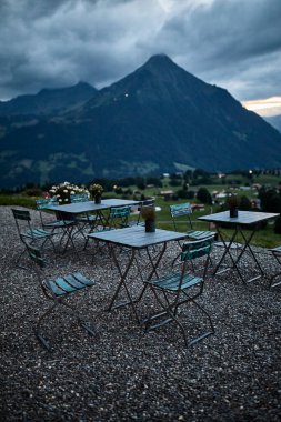 İsviçre Alpleri 'nde bir otelin yanındaki açık hava atıştırma masaları, dağlara bakan bir yer ve sıcak akşam ışığında bir göl. Görüntü rahatlamayı, seyahati, dağlık manzarayı ve akşam yemeğini temsil ediyor. Turizm kavramı, yaz tatili, konukseverlik, rahat
