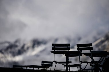 Yemek sandalyeleri İsviçre 'de bir dağ otelinin yanında, sonbahar renklerinde, karla kaplı Alpler' in arka planında. Sahne rahat bir konukseverlik, dağlık bir ortam ve mevsimlik güzelliği temsil ediyor. Seyahat ve açık hava yemekleri için uygun.