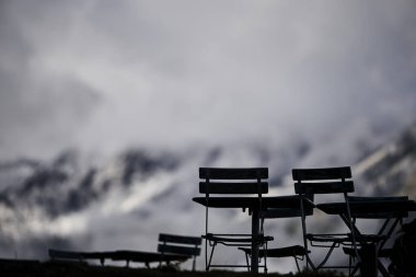 Yemek sandalyeleri İsviçre 'de bir dağ otelinin yanında, sonbahar renklerinde, karla kaplı Alpler' in arka planında. Sahne rahat bir konukseverlik, dağlık bir ortam ve mevsimlik güzelliği temsil ediyor. Seyahat ve açık hava yemekleri için uygun.