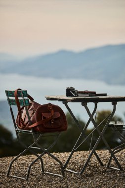 İsviçre 'de bir yaz akşamı manzaralı dağ manzaralı bir terasta sandalye ve masa. Sandalyede sıcak bir eşarp, deri bir seyahat çantası, bir defter ve masanın üzerinde Edelweiss çiçekleri olan bir kamera var. Sahne rahatlatıcı.