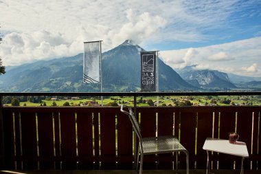 İsviçre yazında dağ manzaralı sandalyeler, çay, kahve ve kitapların yer aldığı bir otel balkonu ve göl manzarası. Sahne, rahat bir dağlık yaşam tarzı, huzurlu bir atmosfer ve rahat bir seyahat deneyimi yakalıyor. Konseptler için uygun