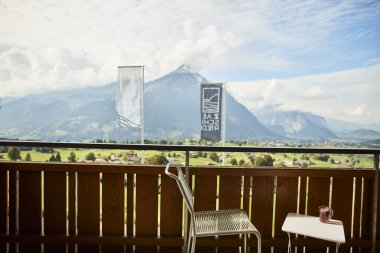 İsviçre yazında dağ manzaralı sandalyeler, çay, kahve ve kitapların yer aldığı bir otel balkonu ve göl manzarası. Sahne, rahat bir dağlık yaşam tarzı, huzurlu bir atmosfer ve rahat bir seyahat deneyimi yakalıyor. Konseptler için uygun