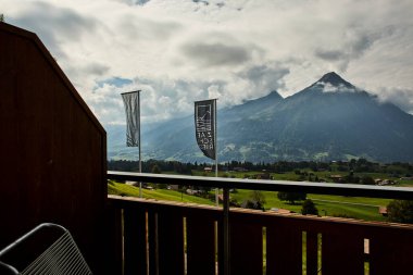 İsviçre yazında dağ manzaralı sandalyeler, çay, kahve ve kitapların yer aldığı bir otel balkonu ve göl manzarası. Sahne, rahat bir dağlık yaşam tarzı, huzurlu bir atmosfer ve rahat bir seyahat deneyimi yakalıyor. Konseptler için uygun