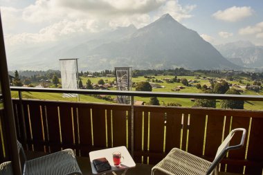 İsviçre yazında dağ manzaralı sandalyeler, çay, kahve ve kitapların yer aldığı bir otel balkonu ve göl manzarası. Sahne, rahat bir dağlık yaşam tarzı, huzurlu bir atmosfer ve rahat bir seyahat deneyimi yakalıyor. Konseptler için uygun