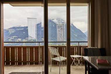 İsviçre yazında dağ manzaralı sandalyeler, çay, kahve ve kitapların yer aldığı bir otel balkonu ve göl manzarası. Sahne, rahat bir dağlık yaşam tarzı, huzurlu bir atmosfer ve rahat bir seyahat deneyimi yakalıyor. Konseptler için uygun