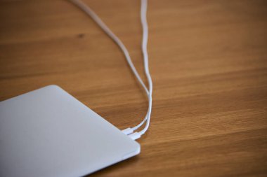 İki USB-C kablosu bağlı, biri şarj için kullanılan ahşap bir masanın üzerinde duran kapalı bir laptop. Sahne, modern bir çalışma alanına, organize masa düzenine ve teknolojiye hazır olmaya dikkat çekiyor. Ofis işleri, üretkenlik ve uzak çalışma kavramları için uygun