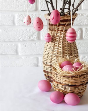 Hasır bir vazoda süslü ağaç dalları ve beyaz arka planda hasır sepette pembe renkli yumurtalar olan Paskalya kompozisyonu. Boşluğu kopyala