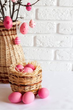 Hasır bir vazoda süslü ağaç dalları ve beyaz arka planda hasır sepette pembe renkli yumurtalar olan Paskalya kompozisyonu. Boşluğu kopyala