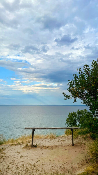 A serene view of the Kaniv Reservoir in Cherkasy region, Ukraine, with a simple wooden bench overlooking the calm waters under a cloudy blue sky.