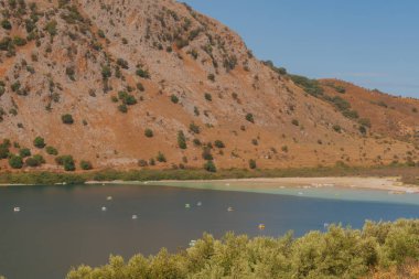 Kuru kıyı şeridi ve turuncu kayalık yamaçlarla kuruyan dağ gölü, sığ sulardaki katamaranlar doğadaki ekolojiyi, turizmi ve kuraklık sorununu simgeliyor.