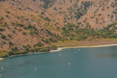 Saklı dağ gölü sakin turkuaz su, kırmızı kayalık yamaç ve yeşil ağaçlar, kumsalda katamaranlar, huzurlu yaz manzarası ve seyahat yeri.