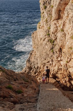 Baba ve kızı macerada: engebeli bir sahil yamacına inen taş basamaklar. Aile bağlarını, seyahatleri, paylaşılan keşifleri ve birlikte geçirilen kaliteli zamanları sembolize eder..