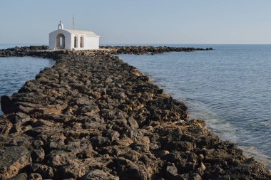 Georgioupolis yakınlarındaki ikonik kıyı şapeli mavi sularla çevrili, açık deniz manzarası boyunca uzun kayalık erişim rotasını ve parlak gündüz kuşağını gösteriyor.