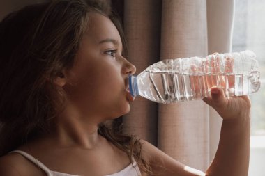 Childs hidrasyonu. Bir kız evin penceresinin yanındaki şişeden su içiyor. Bu görüntü sağlık temalarını, sıvı alımını ve çocukların günlük sağlıklı alışkanlıklarını ve rutinlerini göstermek için mükemmeldir.
