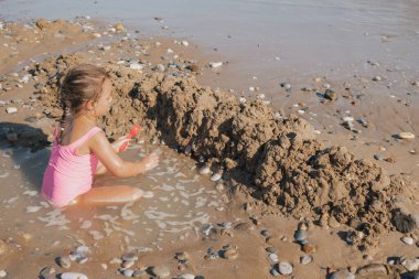 Pembe mayo giymiş küçük bir kız, küçük dalgalarda ve ıslak kumda oynuyor, güneşli bir yaz tatili sırasında çakıl taşı sahilinde kumdan kale inşa ediyor.
