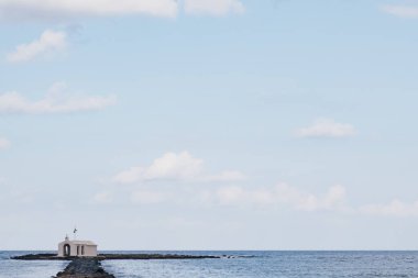 Georgioupolis yakınlarındaki ikonik kıyı şapeli mavi sularla çevrili, açık deniz manzarası boyunca uzun kayalık erişim rotasını ve parlak gündüz kuşağını gösteriyor.