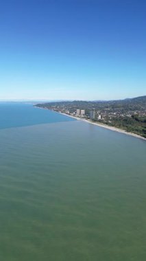 Batumi Botanik Bahçesi yakınlarındaki Karadeniz kıyısı. Dikey 4K drone görüntüleri yemyeşil yemyeşil, kıyı kayalıkları ve açık güneşli havada açık mavi deniz manzaraları gösteriyor..