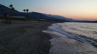 Gün batımında Girit 'te palmiyeler ve dağlarla dalgalar. 4K drone videosu, sakin bir Akdeniz atmosferinde altın akşam ışığı, dalgalı deniz dalgaları, tropikal palmiyeler ve uzak dağ siluetini yakalıyor.