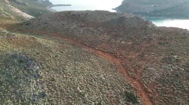Girit 'teki uzak Balos Körfezi' ne giden toprak yol. İnsansız hava aracı görüntüleri engebeli Yunan manzarası, yürüyüş rotası, dik arazi ve ulaşılması zor kıyı lagününün yakınındaki turkuaz Akdeniz suyunu gösteriyor.