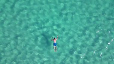İHA 'nın yukarıdan görünüşü: Turkuaz deniz suyunda yüzen adam baş parmak hareketi gösteriyor, Elafonisi pembe kumsal.
