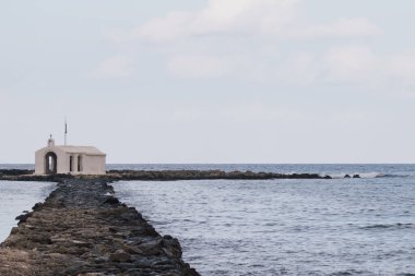 Parlak gün ışığıyla çerçevelenmiş kıyı manzaralı, Georgioupolis 'in beyaz deniz kenarı kilisesi, uzun taş erişim yolu ve binanın etrafındaki yumuşak su yüzeyi.