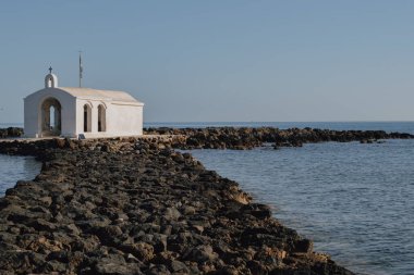 Georgioupolis yakınlarındaki ikonik kıyı şapeli mavi sularla çevrili, açık deniz manzarası boyunca uzun kayalık erişim rotasını ve parlak gündüz kuşağını gösteriyor.