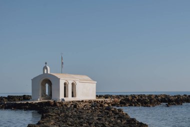 Parlak gün ışığıyla çerçevelenmiş kıyı manzaralı, Georgioupolis 'in beyaz deniz kenarı kilisesi, uzun taş erişim yolu ve binanın etrafındaki yumuşak su yüzeyi.
