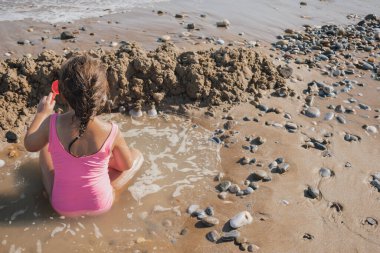 Pembe mayo giymiş küçük bir kız, küçük dalgalarda ve ıslak kumda oynuyor, güneşli bir yaz tatili sırasında çakıl taşı sahilinde kumdan kale inşa ediyor.