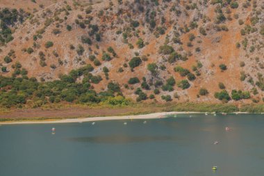 Kuru kıyı şeridi ve turuncu kayalık yamaçlarla kuruyan dağ gölü, sığ sulardaki katamaranlar doğadaki ekolojiyi, turizmi ve kuraklık sorununu simgeliyor.