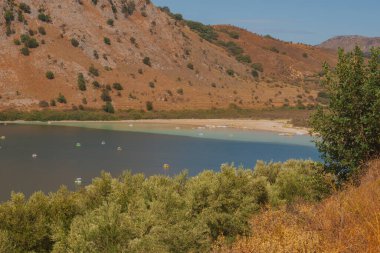 Kuru kıyı şeridi ve turuncu kayalık yamaçlarla kuruyan dağ gölü, sığ sulardaki katamaranlar doğadaki ekolojiyi, turizmi ve kuraklık sorununu simgeliyor.