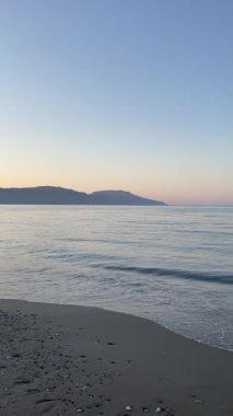 Dikey çekim: küçük deniz dalgaları ve gün doğumunda uzak dağ siluetiyle kumlu sahil üzerinde yavaş uçuş