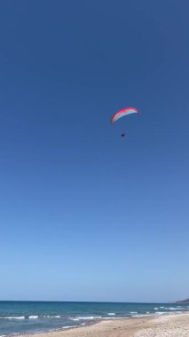Deniz ve sahil üzerinde yamaç paraşütü yapan erkek pilotun dikey videosu, güneşli hava, Yunanistan 'da yaz tatilinde aşırı turistik etkinlik.