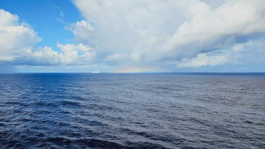 Uzak yolcu gemisi ve ender görülen yatay gökkuşağı ile geniş deniz manzarası. Derin mavi su dalgalanmaları ve değişen gökyüzünün altında dinamik kümülüs bulutları..