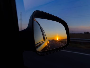 Sıcak renkleri, açık yol manzarasını ve sakin bir akşam atmosferini gösteren araba yan aynasında altın bir günbatımı..